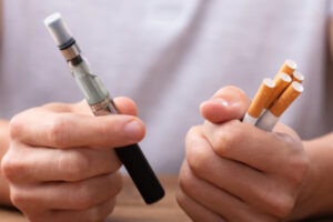 Is Vaping Worse than Smoking? A close up shot of a vaping pen in one hand and five cigarettes in the other.
