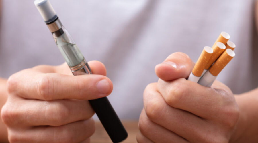 Is Vaping Worse than Smoking? A close up shot of a vaping pen in one hand and five cigarettes in the other.